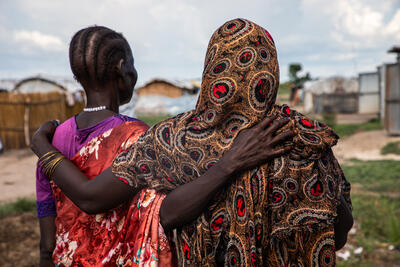 Two women stand in a open space with their backs to the camera. They have an arm around each other's back.