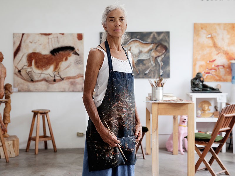 Female artist holding a paint brush standing in front of paintings