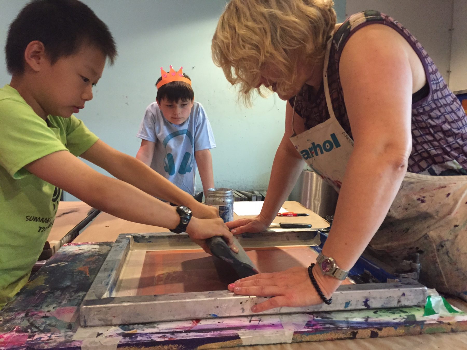 A blonde woman wearing a Warhol apron helps a young boy in a green t shirt make a screen print as another young boy wearing an orange paper crown leans forward to watch.