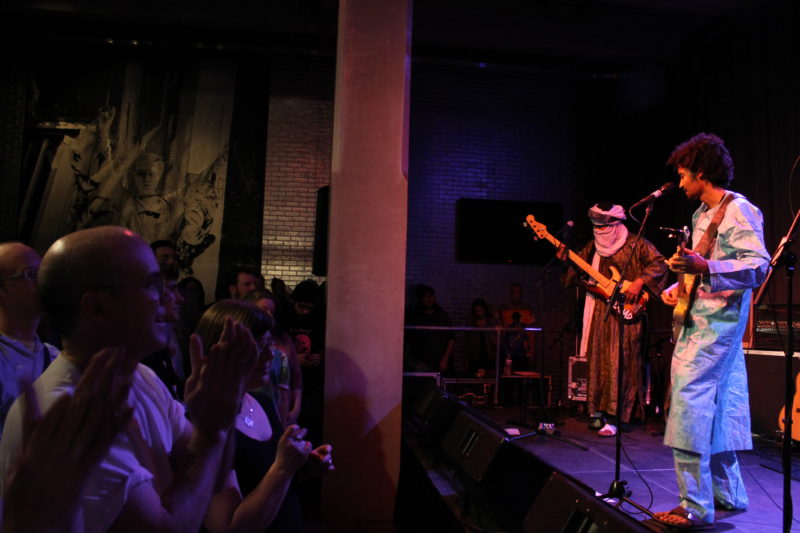 Two performers stand onstage to the right side of the image. On the left, a crowd claps.