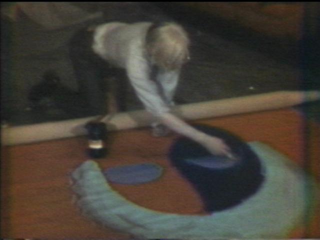 A man kneeling over the edge of a large canvas painting blue eye shadow onto a portrait