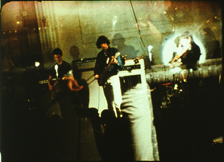 A grainy photograph of two guitar players and a violinist, with the figure of the photographer reflected in a nearby window