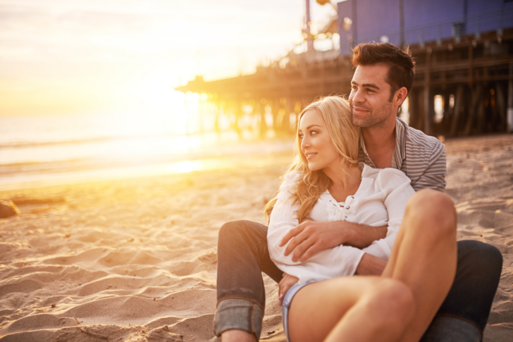 Ein Mann umarmt eine Frau von hinten, während sie glücklich am Strand sitzen und den Sonnenuntergang beobachten.