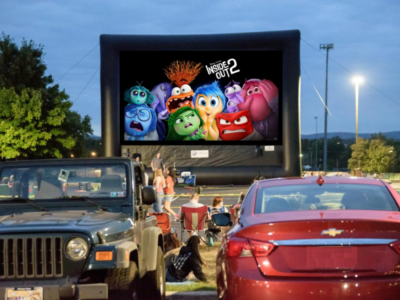 Cars face a large inflatable movie screen for a drive-in movie.