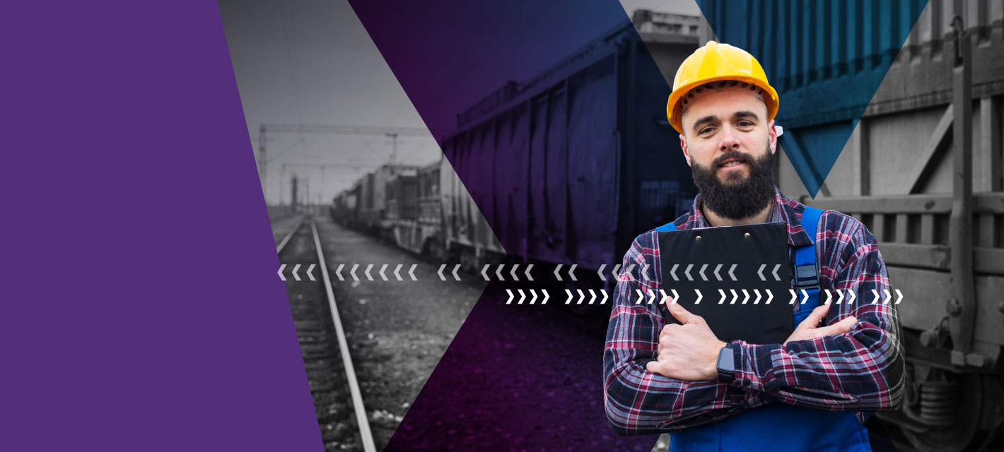 A man wearing a hard hat holds a tablet while standing beside a freight train in a rail yard.